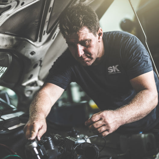 A mechanic works on an engine under a car hood, focused on repairing machinery.